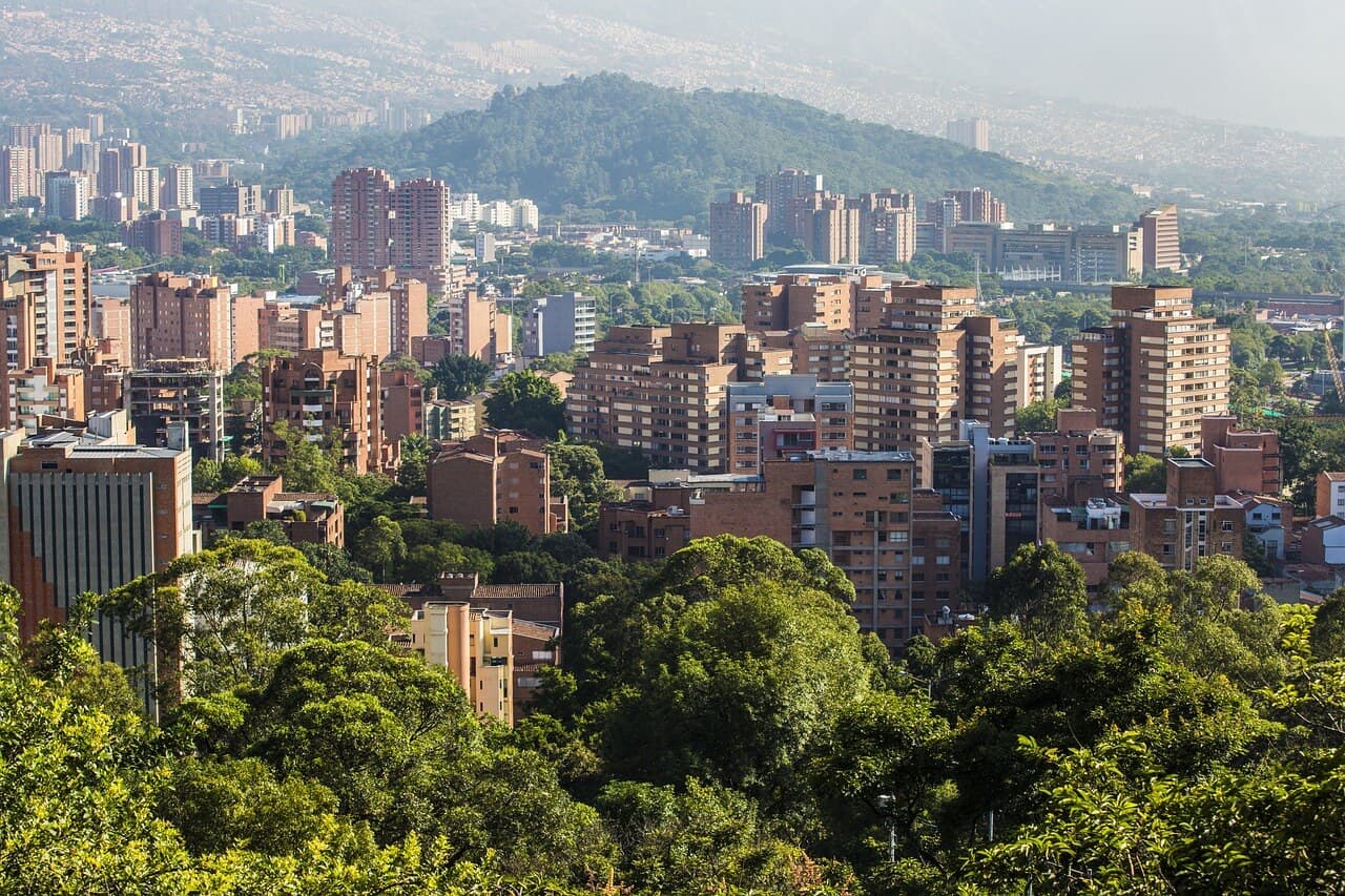 A sunny daytime view of Medellín city, featuring buildings and lush green trees surrounded by mountains.